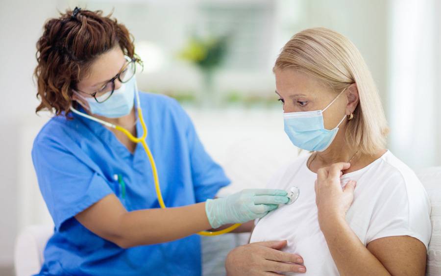 A woman with a surgical mask holds her chest while a medical professional listens to her lungs with a stethoscope.