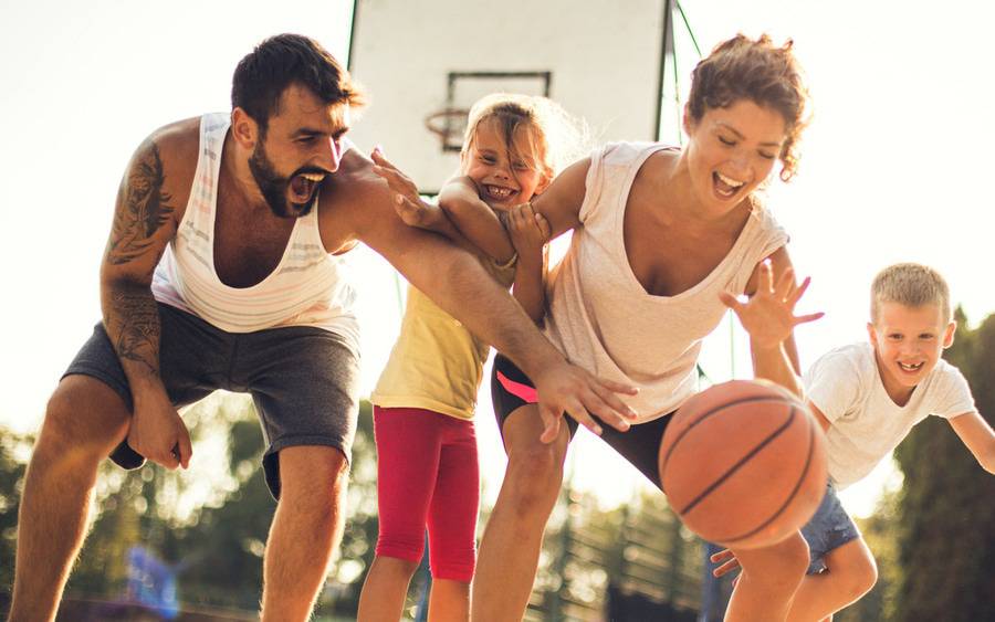 A young family smiles as they reach for the basketball during a game outdoors.