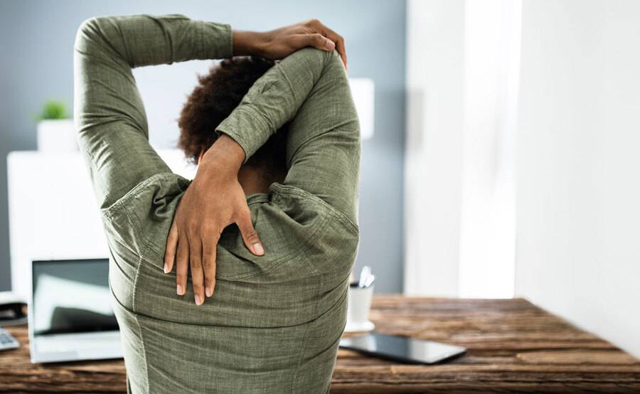 A man at a desk reaches his arm behind his head to stretch his back and neck.