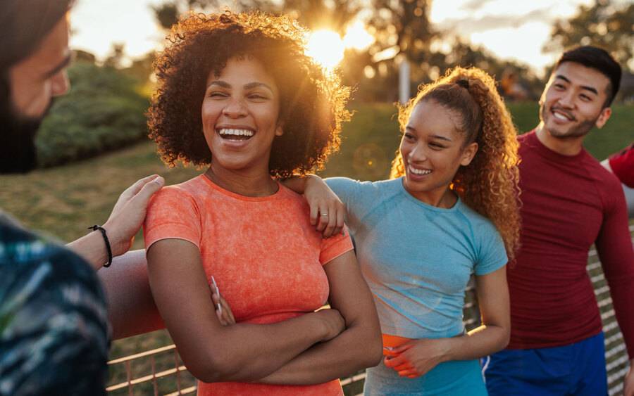 A group of young men and women wearing fitness outfits smile as they gather to do an outdoor workout.