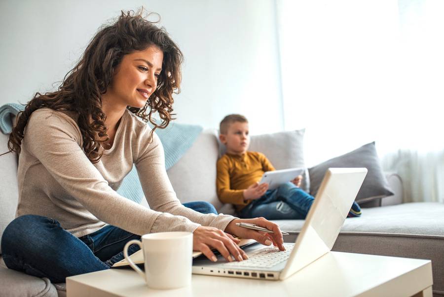 A woman types on a laptop, while a young  boy plays on a table in the background.