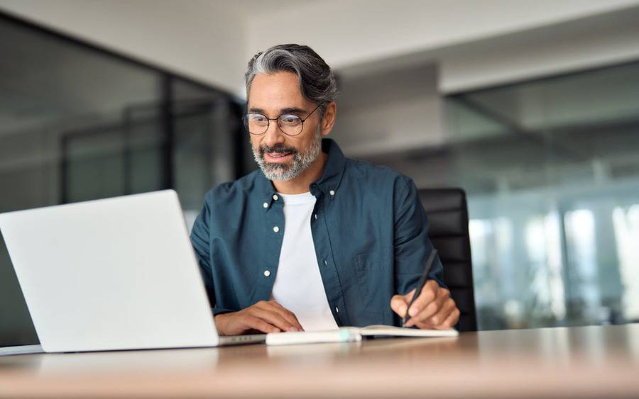 A middle aged man in glasses takes notes while looking at his laptop.