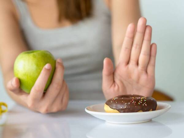 Woman using one hand  to resist eating a doughnut while holding a healthy apple that she plans to eat.