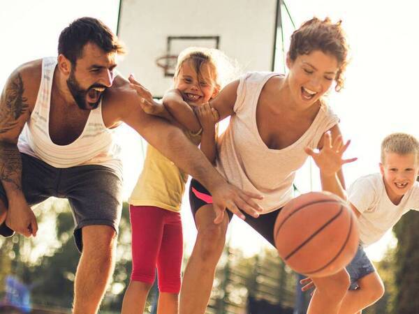 A young family smiles as they reach for the basketball during a game outdoors.